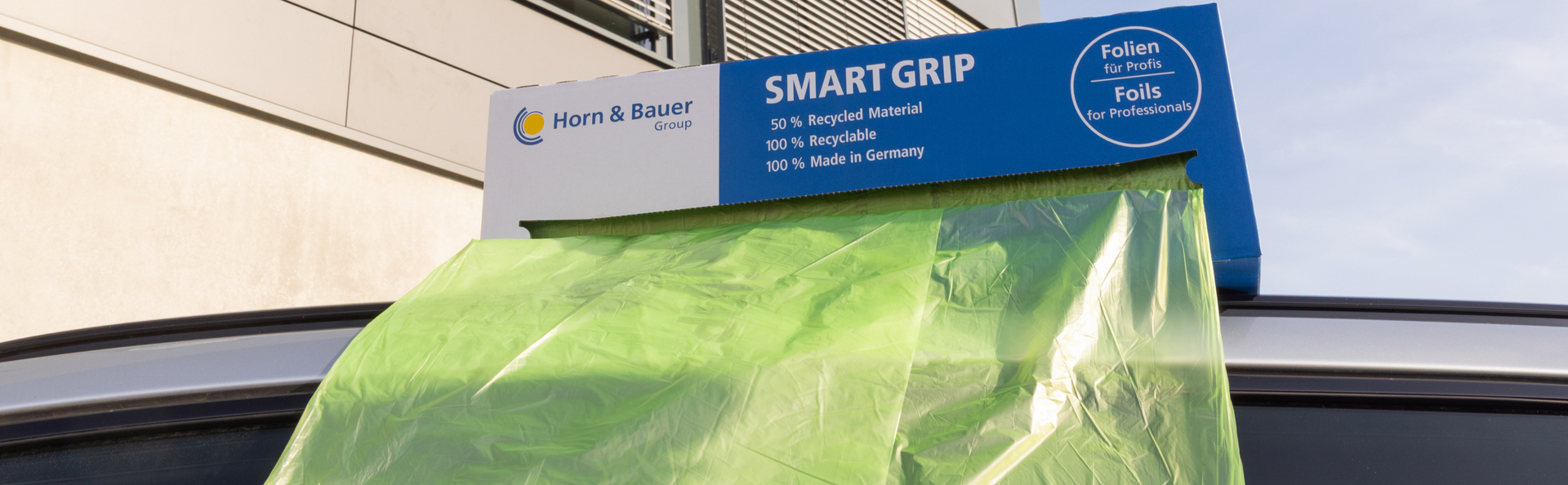 The image shows blue and white packaging from the Horn & Bauer Group brand bearing the inscription ‘SMART GRIP’. Beneath this it reads: ‘50% recycled material, 100% recyclable, 100% made in Germany’. On the right is a circle with the text ‘Folien für Profis / Foils for Professionals’. The Smartgrip3in1 Recycled is being pulled out of the packaging and is hanging over the roof of a vehicle. Parts of a building and the sky can be seen in the background.