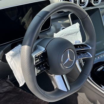The picture shows the steering wheel of a Mercedes-Benz in the car’s interior. The steering wheel is black with silver accents and features the Mercedes logo in the centre. There are controls on the spokes, and some parts are still covered with the Smartgrip3in1 Eco. Parts of the dashboard and a display are visible in the background.